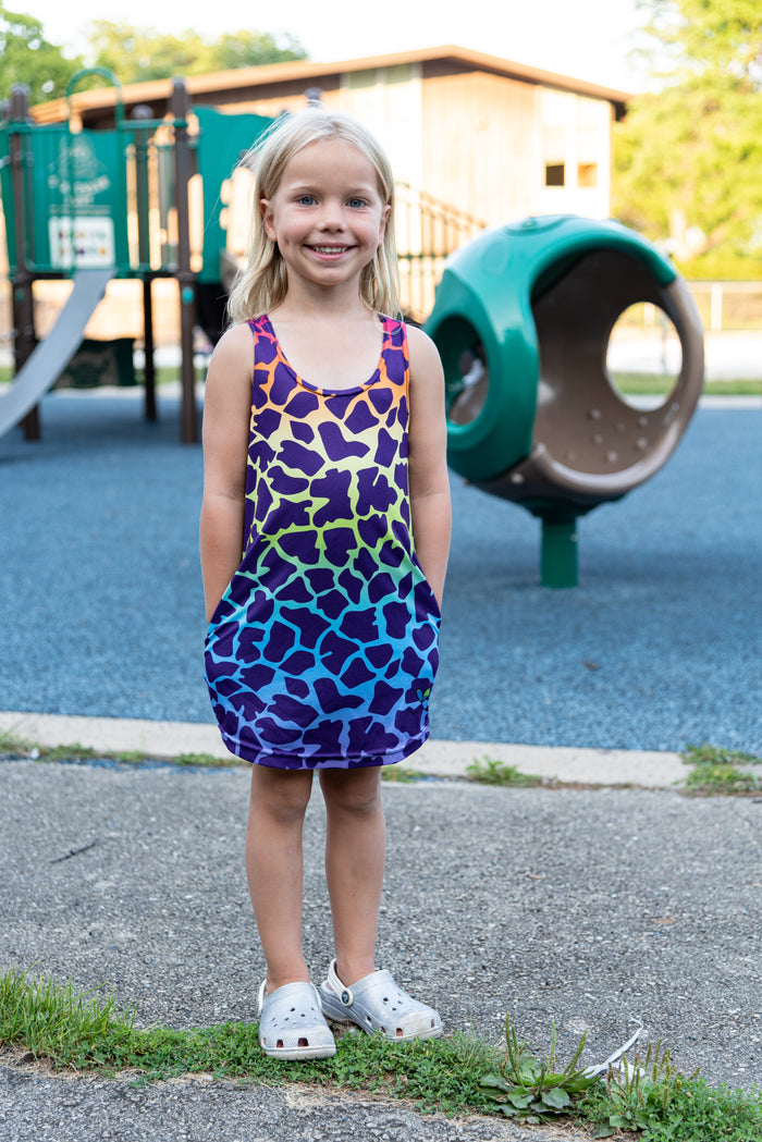 A sweet little blond girl smiling wearing a rainbow giraffe print dress with her hands in her pockets.